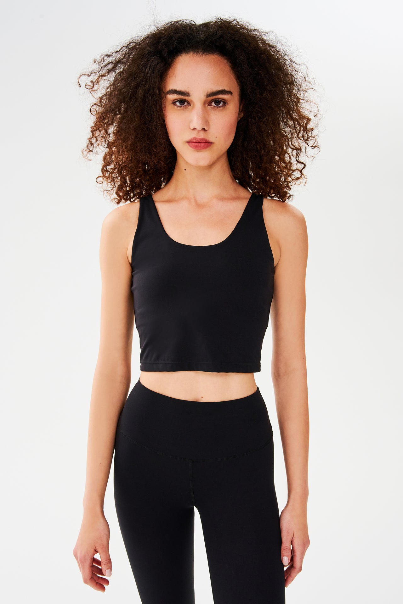 Front view of woman with curly dark brown hair wearing a black bra crop tank with black leggings