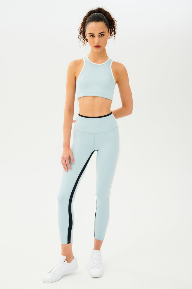 Full front view of girl wearing light blue sports bra with white hem and light blue leggings with black stripe around the waistband and white stripe down the outer seam and a black stripe on inseam and white shoes