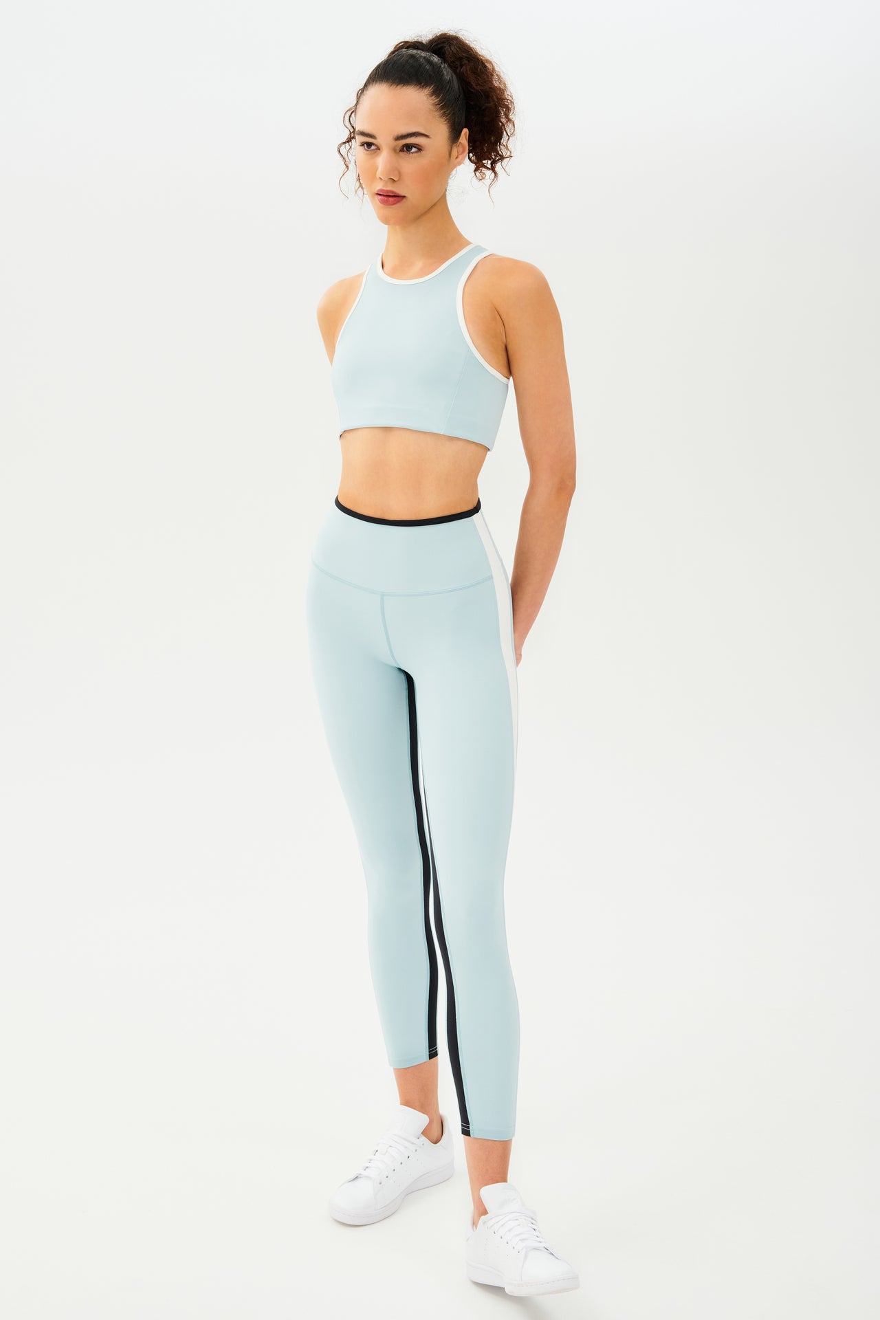 Full side view of girl wearing light blue sports bra with white hem and light blue leggings with black stripe around the waistband and white stripe down the outer seam and a black stripe on inseam and white shoes