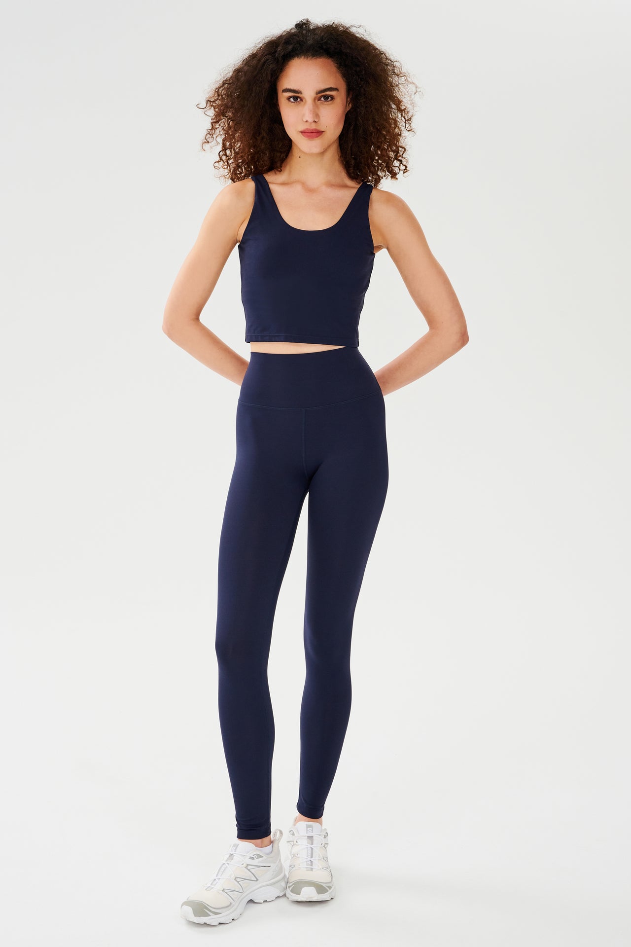 Front full view of woman with dark curly hair wearing dark blue high waist leggings and dark blue bra tank paired with white shoes 