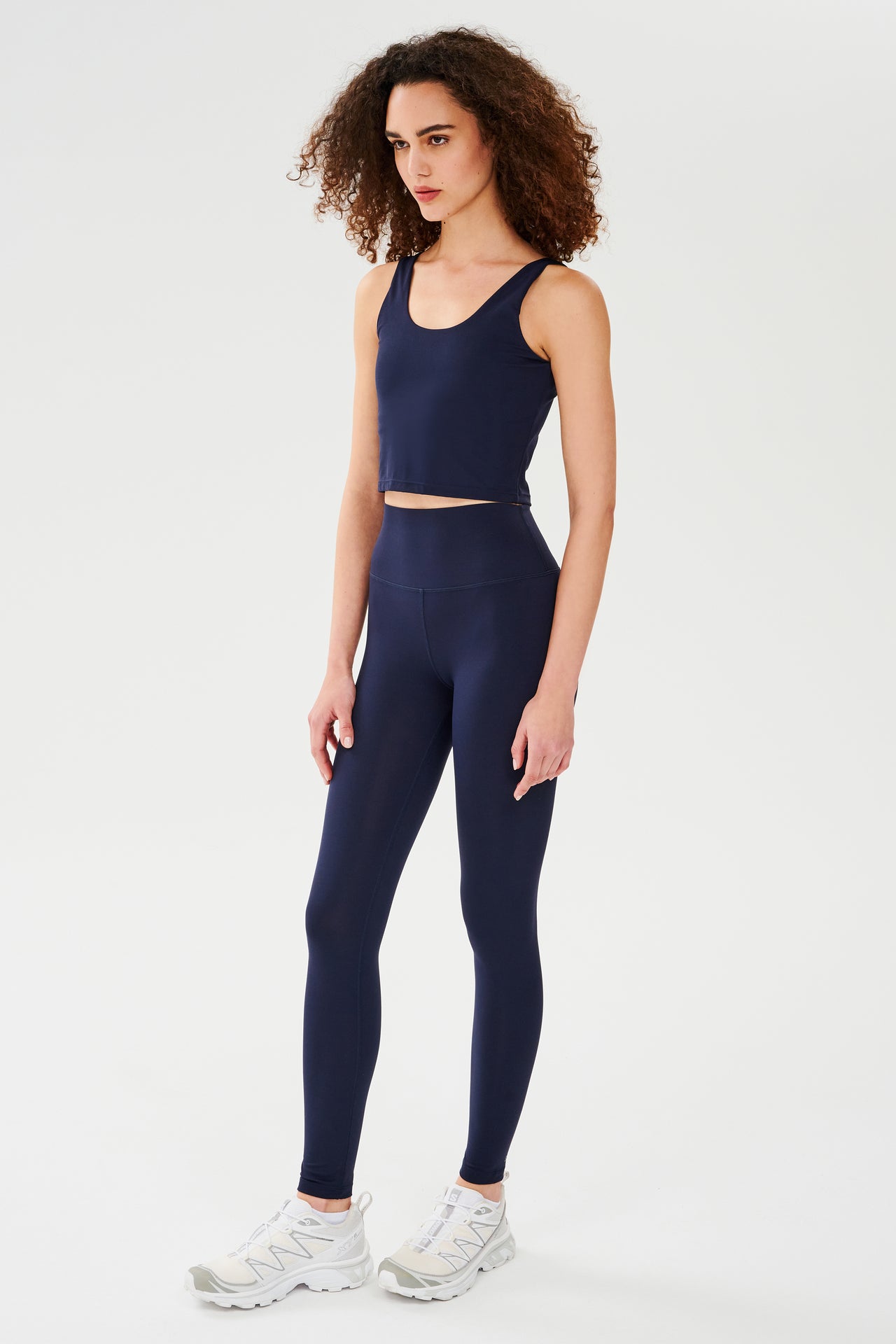 Front full side view of woman with dark curly hair wearing dark blue high waist  leggings and dark blue bra tank paired with white shoes 