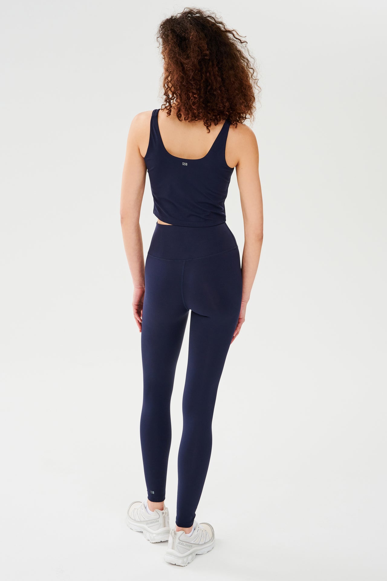 Back full view of woman with curly dark brown hair wearing a dark blue bra crop tank with dark blue leggings and white shoes  