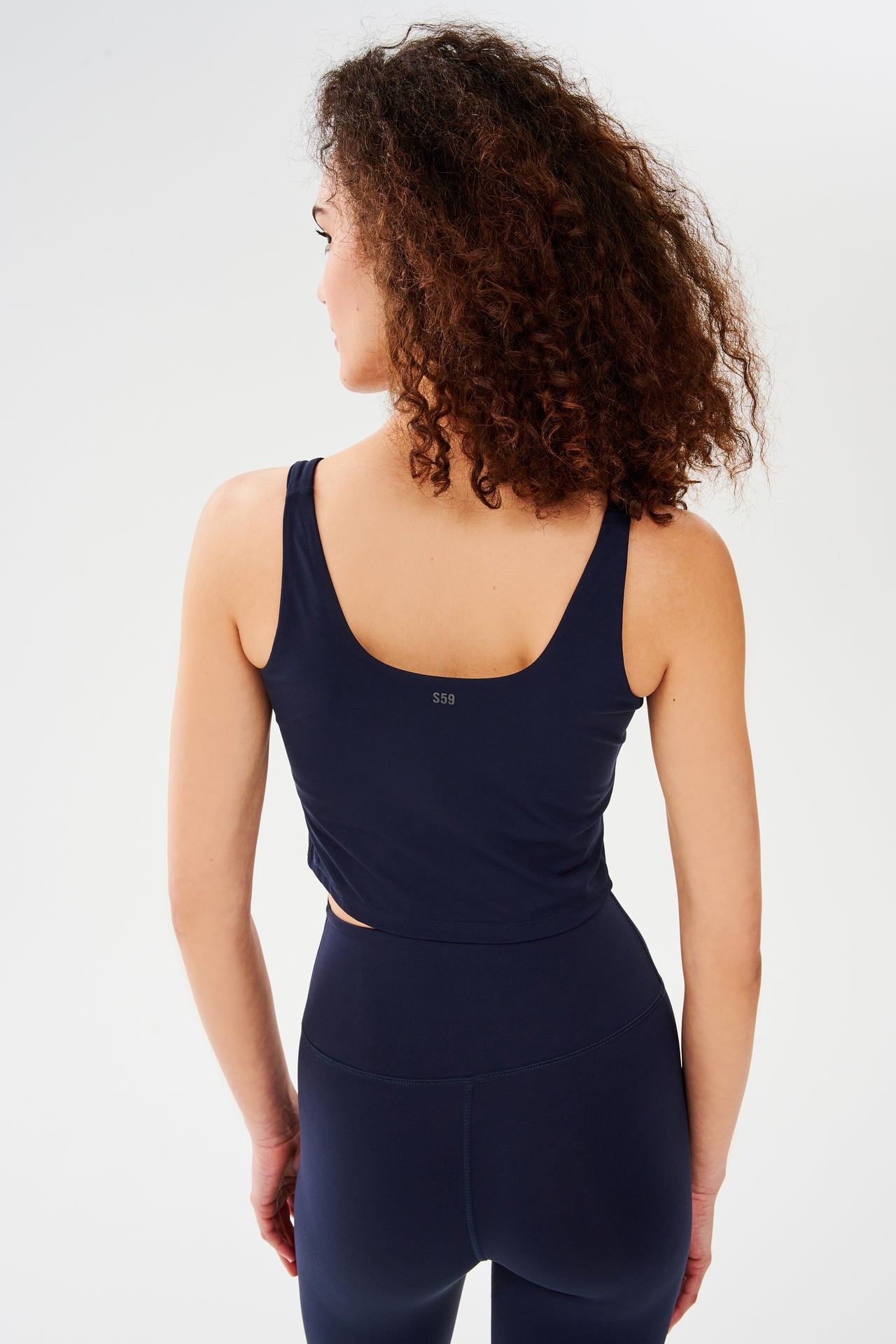 Back view of woman with curly dark brown hair wearing a dark blue bra crop tank with dark blue leggings 
