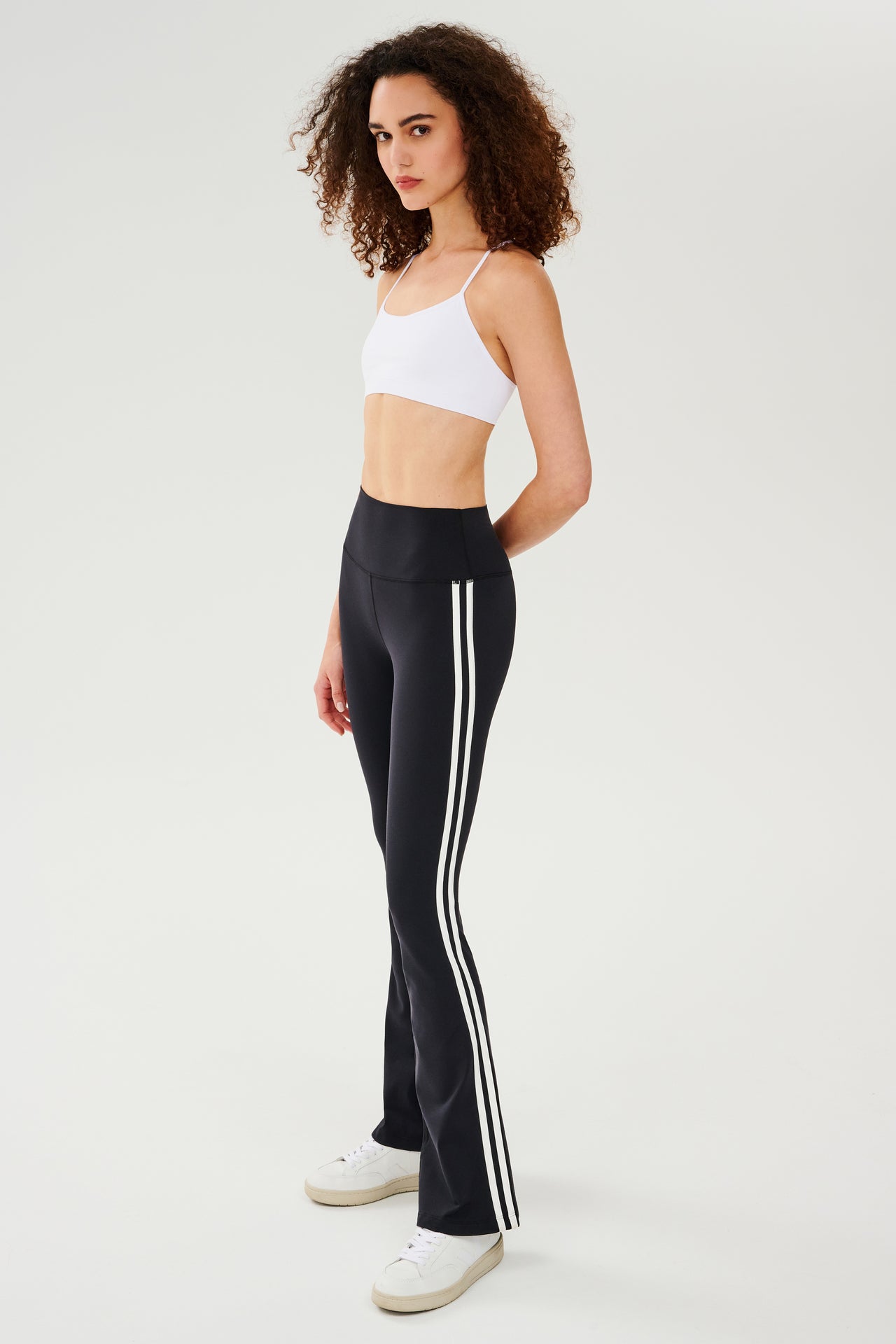 Full front side view of woman with dark curly hair wearing black high waist below ankle length legging with wide flared bottoms with double white side stripes on both legs. Also wearing a white racerback bra with spaghetti straps. Paired with white shoes.