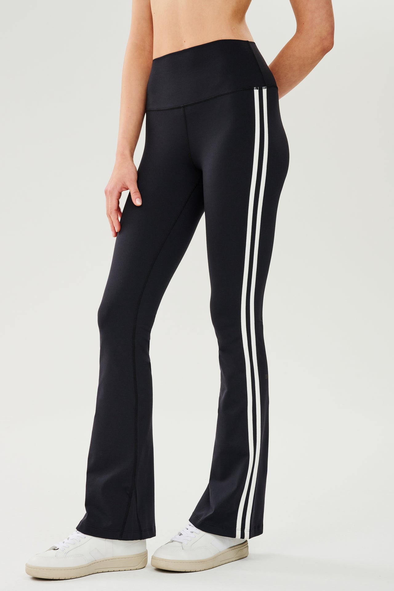 Front view of woman wearing black high waist below ankle length legging with wide flared bottoms with double white side stripes on both legs.Paired with white shoes.