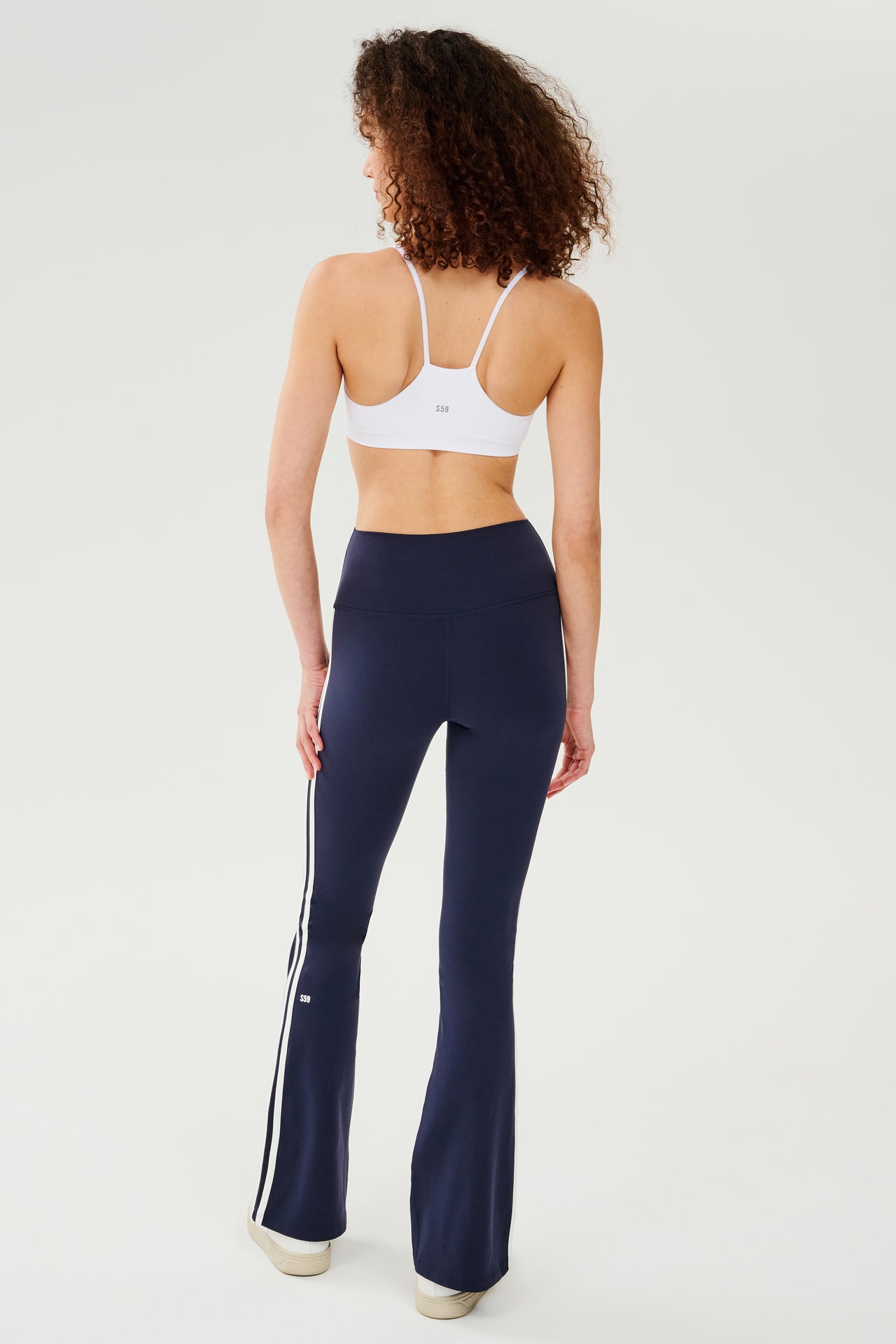 Full back view of woman with dark curly hair wearing dark blue high waist below ankle length legging with wide flared bottoms and white double side stripes and white racerback bra with spaghetti straps. Paired with white shoes. 