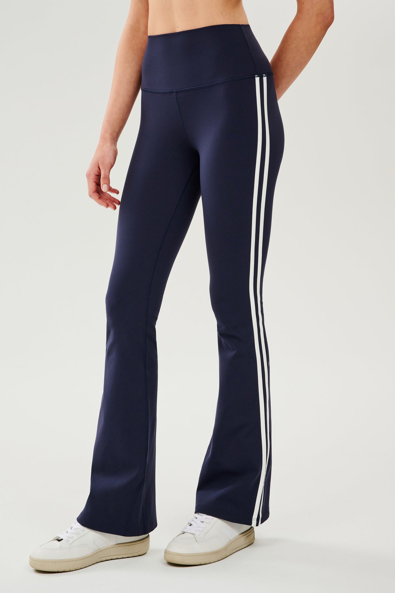 Front view of woman wearing dark blue high waist below ankle length legging with wide flared bottoms and white double side stripes on both legs. Paired with white shoes. 