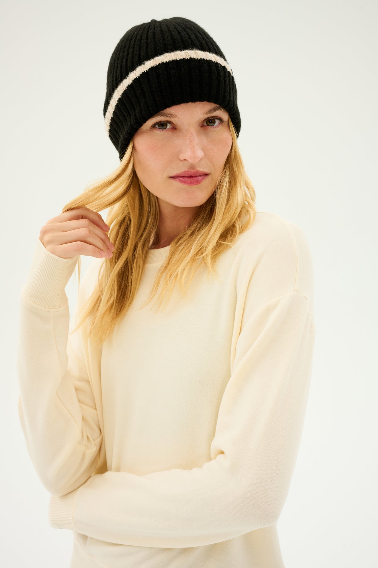 A person with long blonde hair, wearing the SPLITS59 Rib Beanie in Black/Creme and a cozy cream-colored sweater, looks directly at the camera.