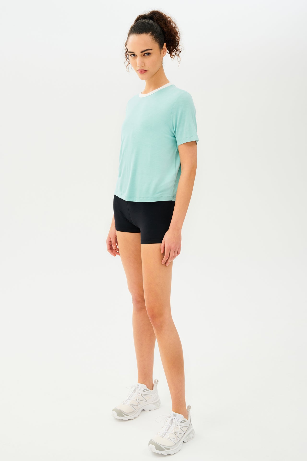 Full side view of girl wearing light blue cropped short sleeve t-shirt with thin white neck hem and black bike shorts with white shoes