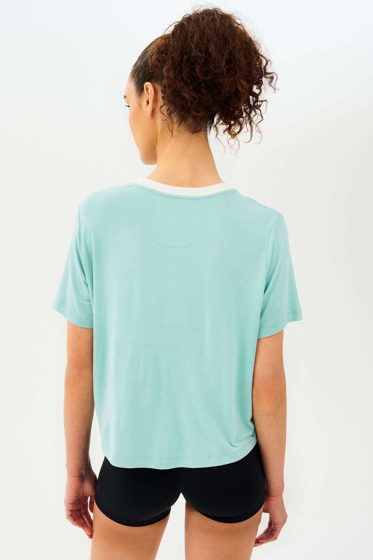 Back view of girl wearing light blue cropped short sleeve t-shirt with thin white neck hem and black bike shorts 