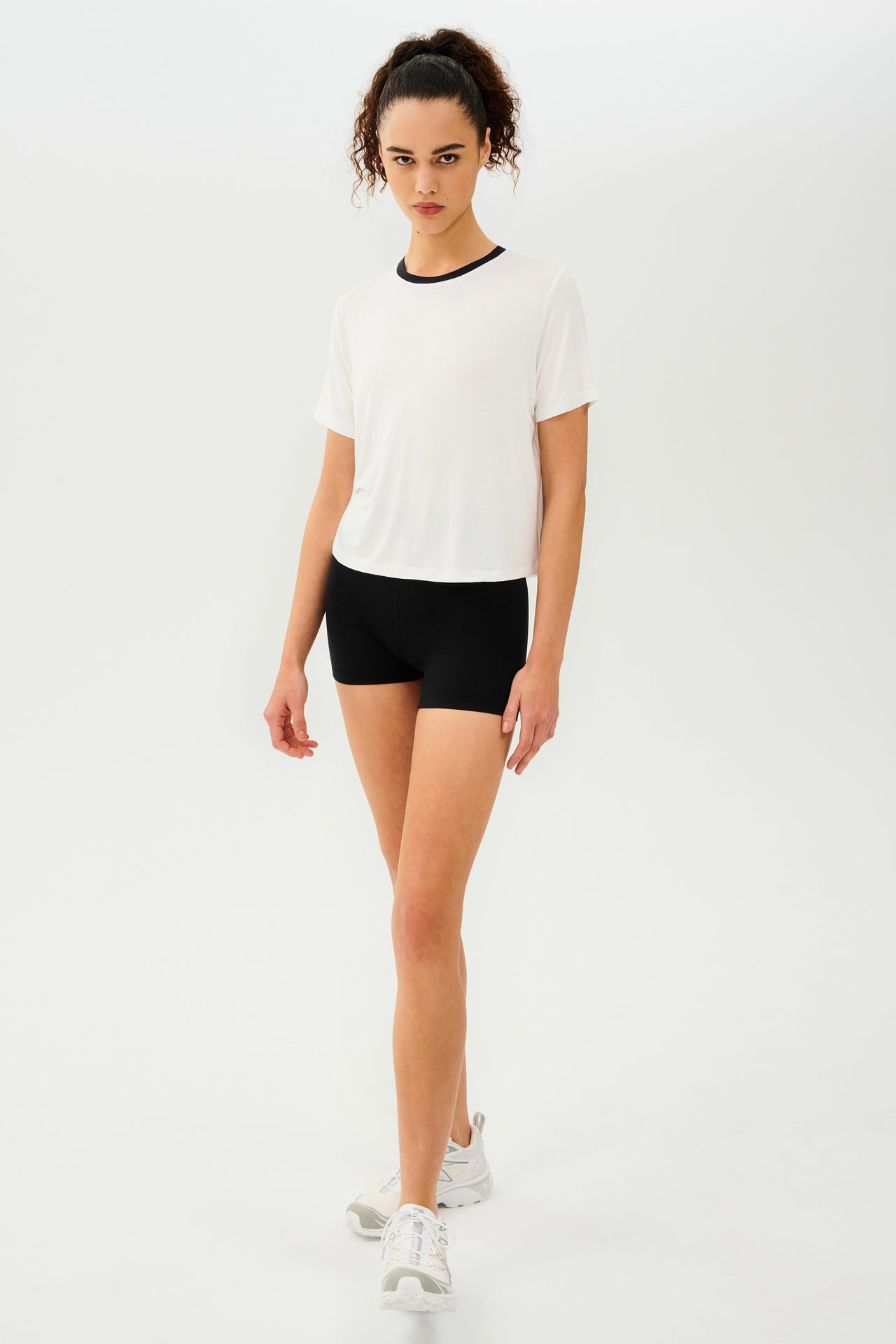 Full side view of girl wearing white cropped short sleeve t-shirt with thin black neck hem and black bike shorts with white shoes