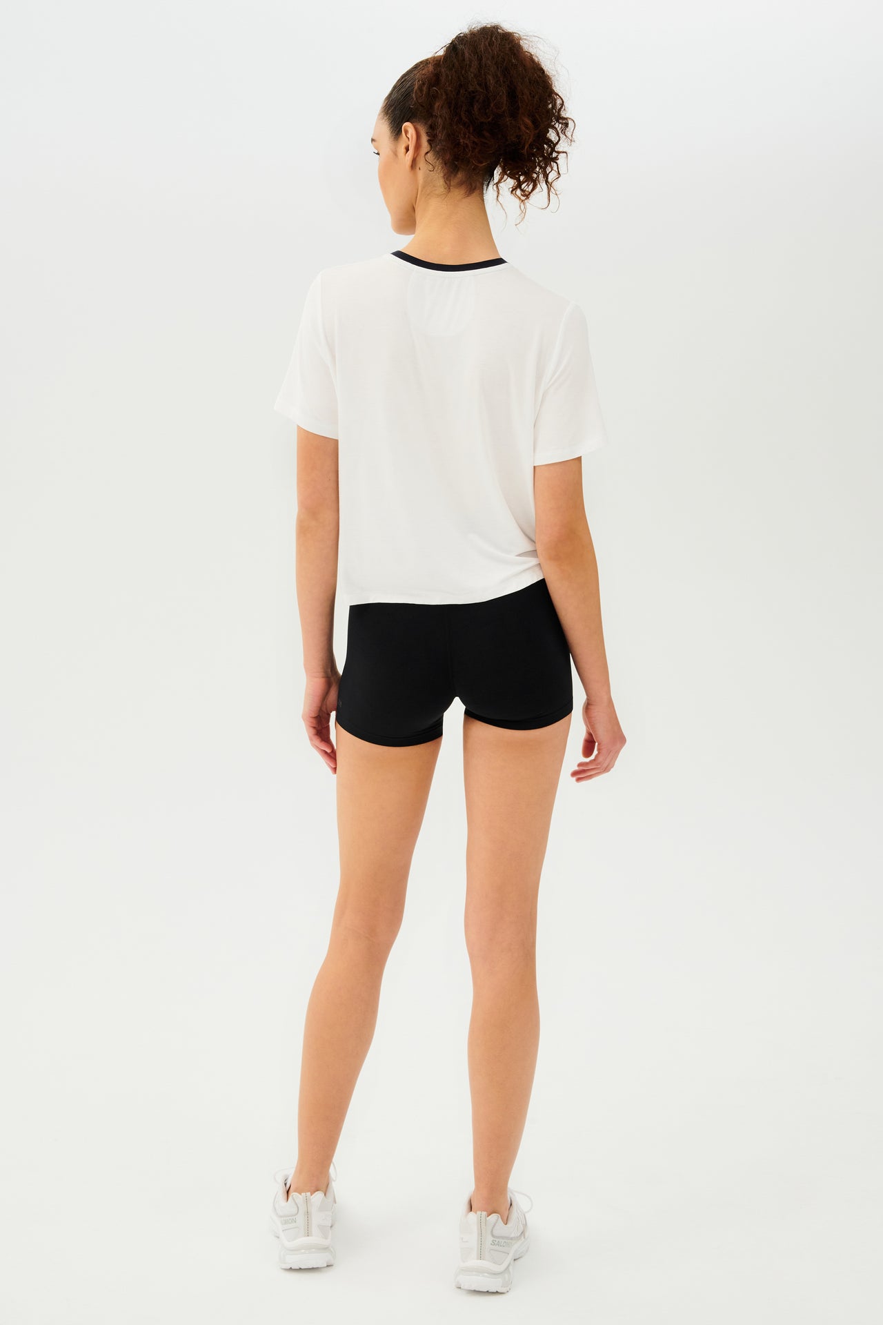 Full back view of girl wearing white cropped short sleeve t-shirt with thin black neck hem and black bike shorts with white shoes