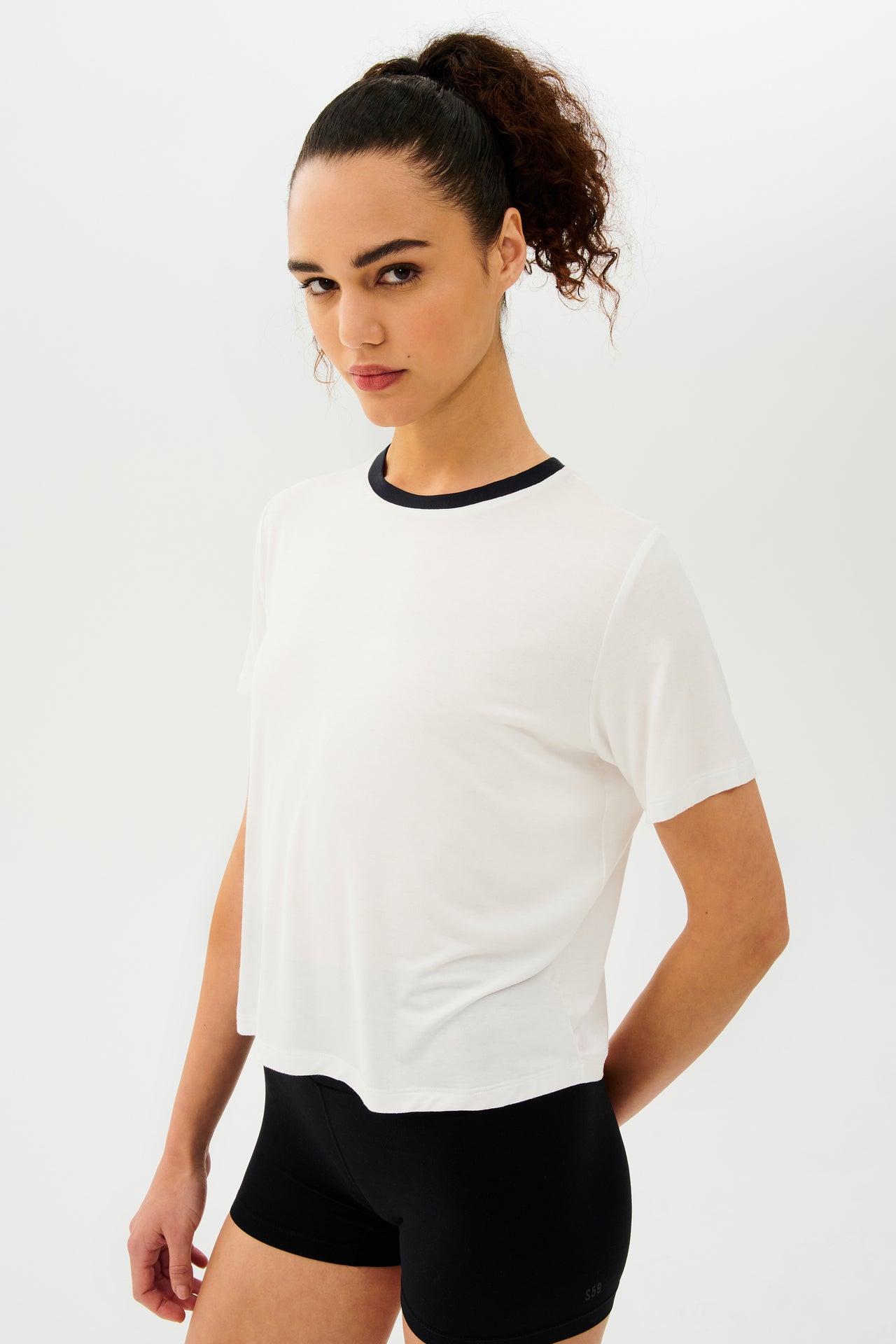 Side view of girl wearing white cropped short sleeve t-shirt with thin black neck hem and black bike shorts 