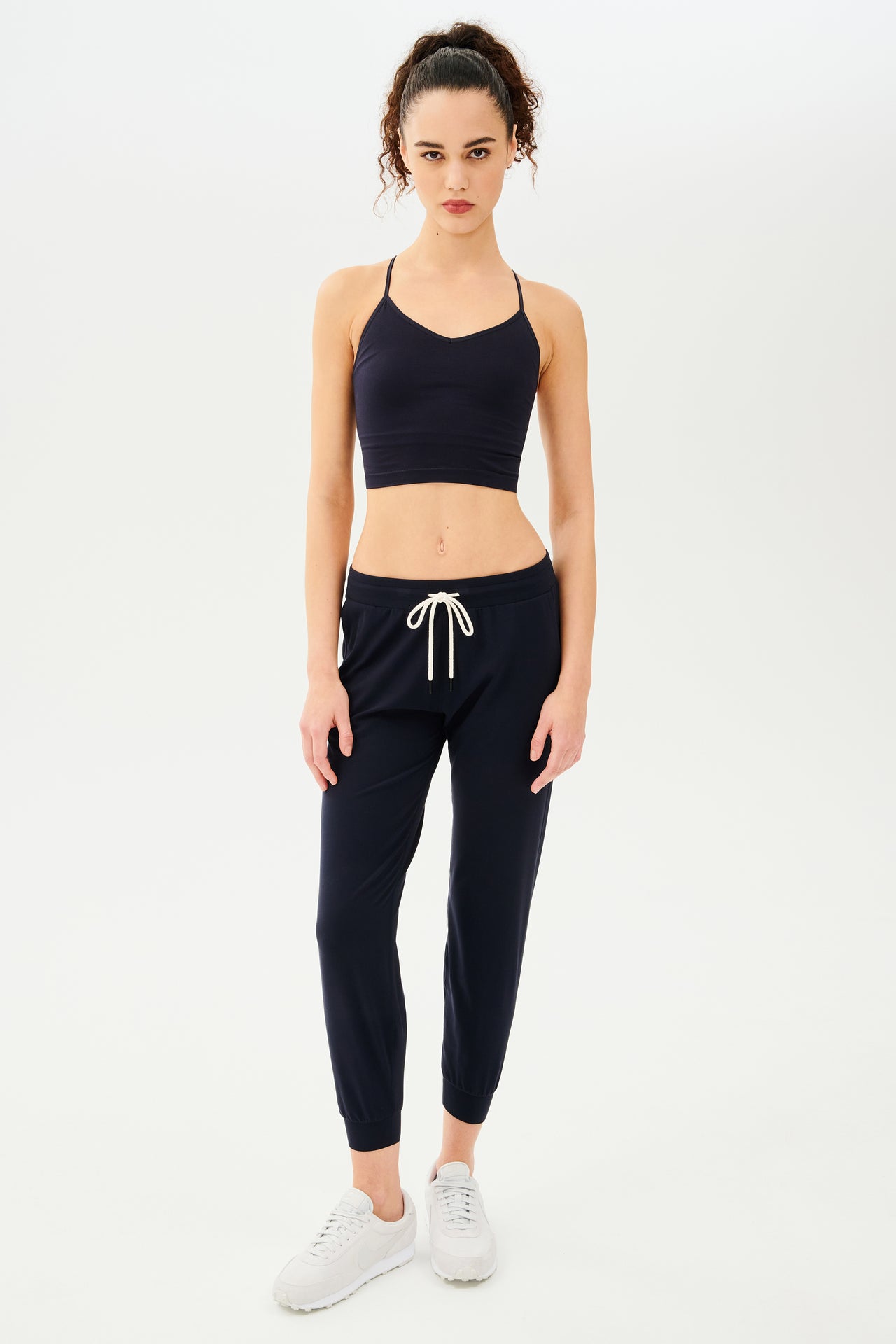 The model is wearing a Splits59 Loren Seamless Cami - Indigo with a built-in shelf bra and black joggers, made from chafe-free fabric.