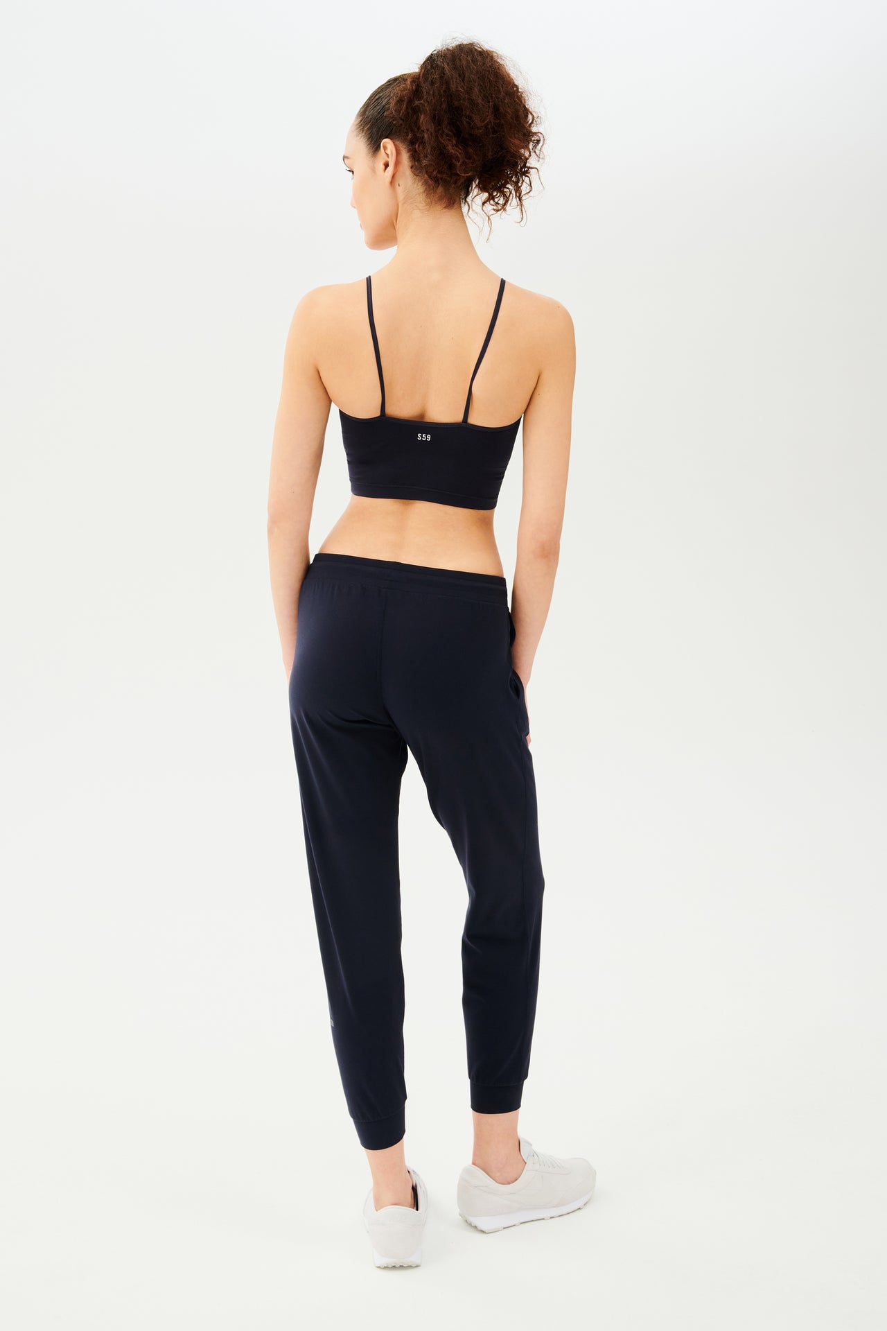 Full back view of girl wearing dark blue sweatpants with black tie around waistband and a dark blue sports bra with white shoes 