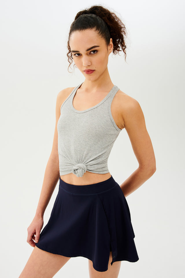 Front full view of woman with dark curly hair in a ponytail wearing a grey tank top and dark blue skort