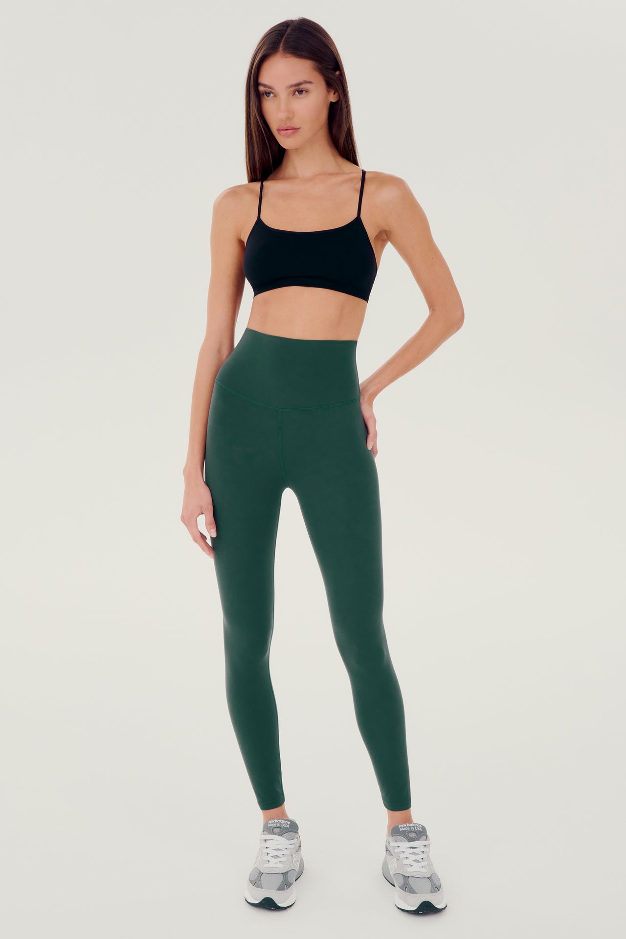 Full front view of girl wearing dark green leggings right above the ankle with black sports bra and grey shoes