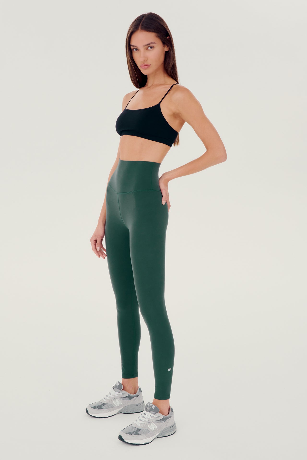 Full side view of girl wearing dark green leggings right above the ankle with black sports bra and grey shoes
