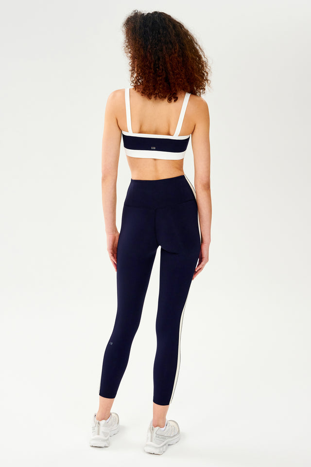 Full back view of girl wearing dark blue leggings with a white down the side and a dark blue and white sports bra with white shoes 