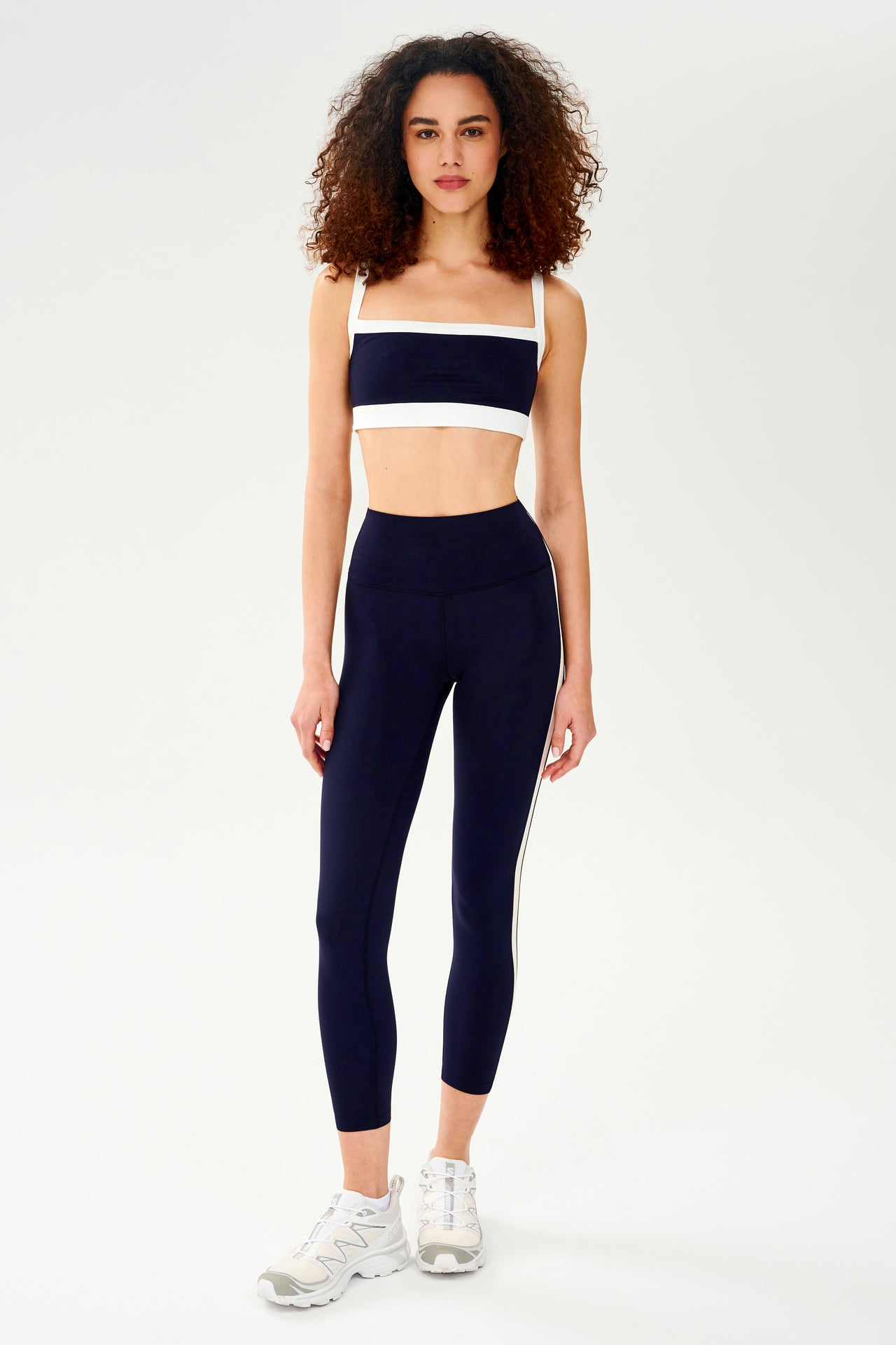 Full front view of girl wearing dark blue leggings with a white down the side and a dark blue and white sports bra with white shoes 