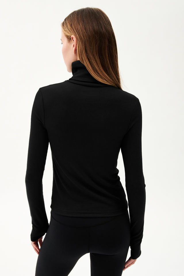 The back view of a woman wearing SPLITS59's Jackson Rib Full Length Turtleneck in Black yoga leggings and a turtleneck top.