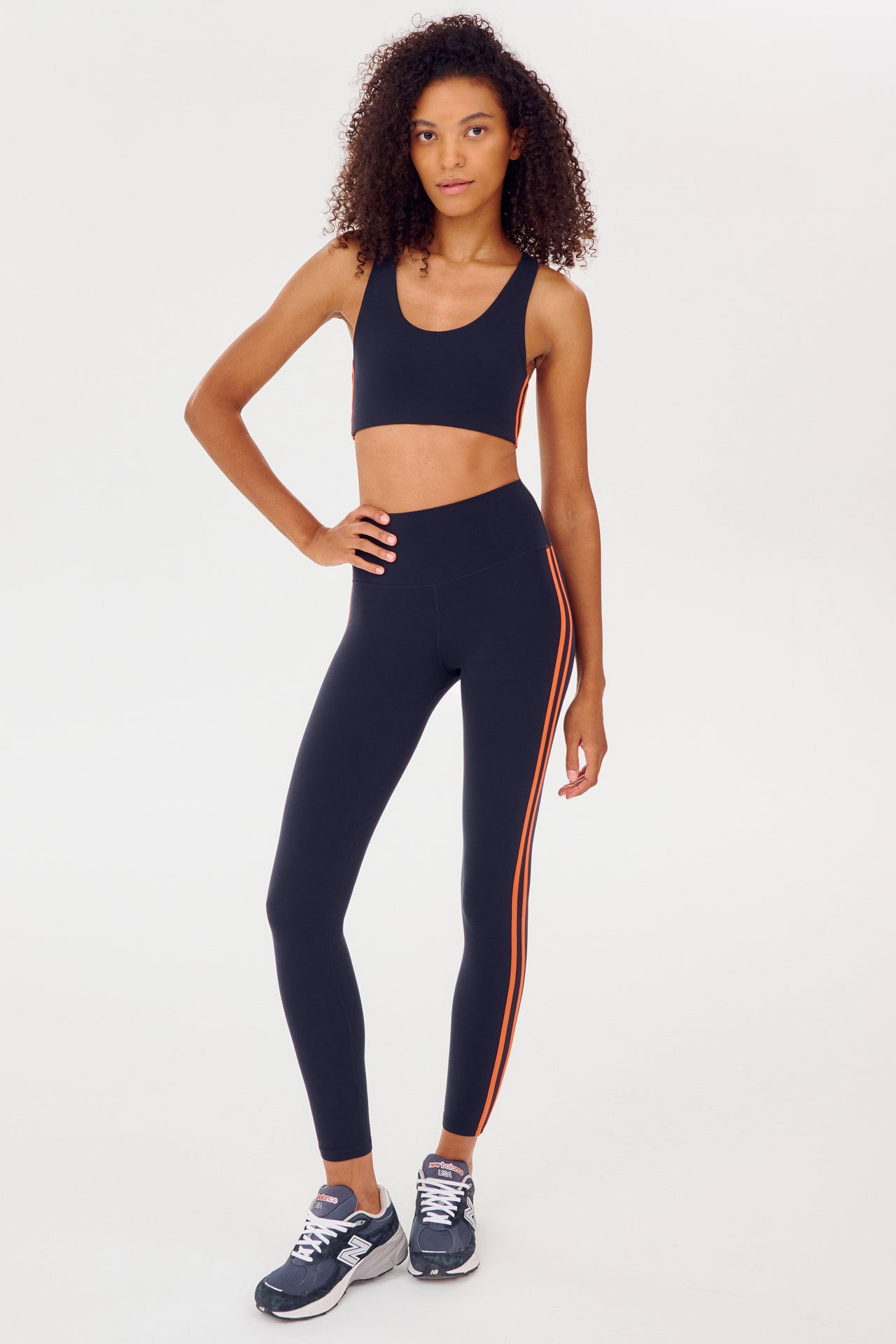 Full front view of girl wearing dark blue leggings with two thin red stripes down the side and a dark blue sports bra with dark blue shoes