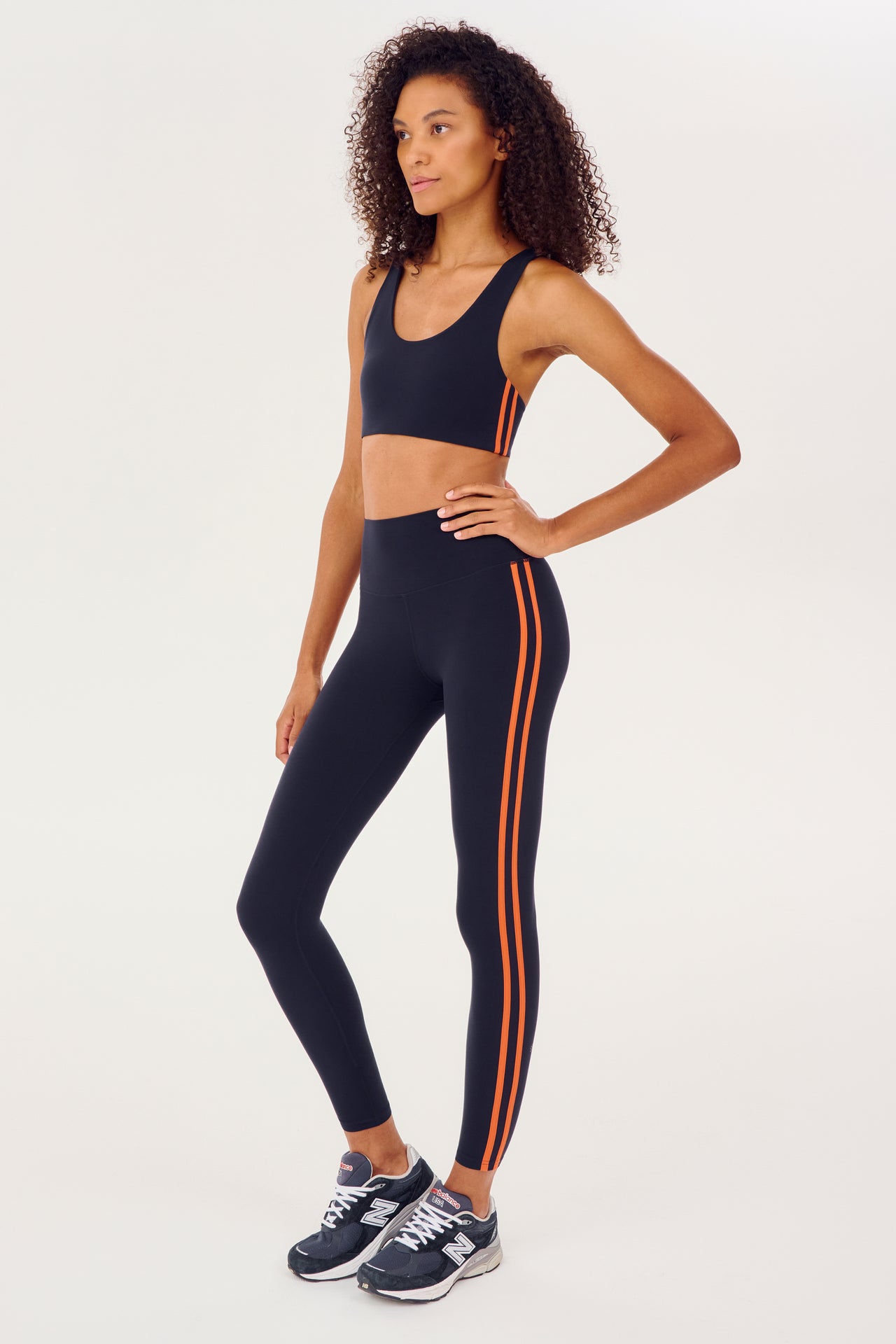 Full side view of girl wearing dark blue leggings with two thin red stripes down the side and a dark blue sports bra with dark blue shoes