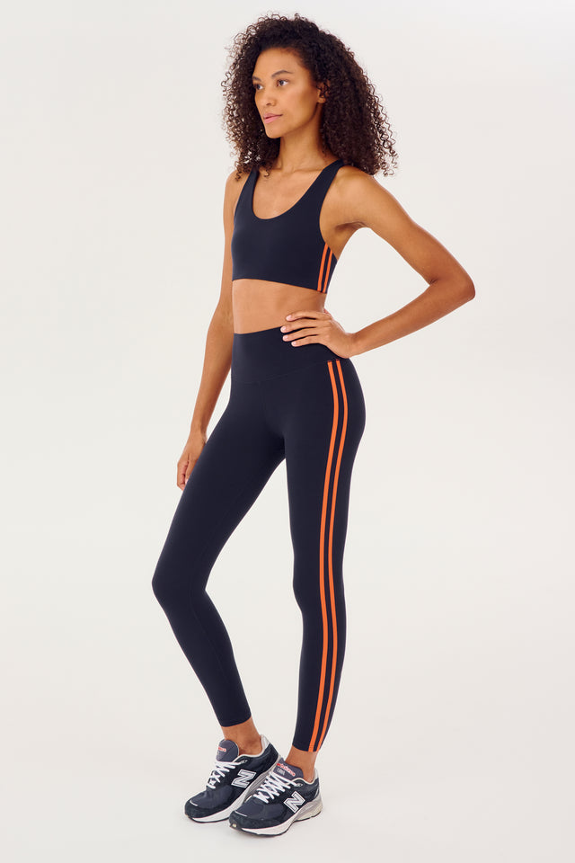 Full side view of girl wearing dark blue sports bra with two thin red stripes down the side and dark blue leggings with dark blue shoes