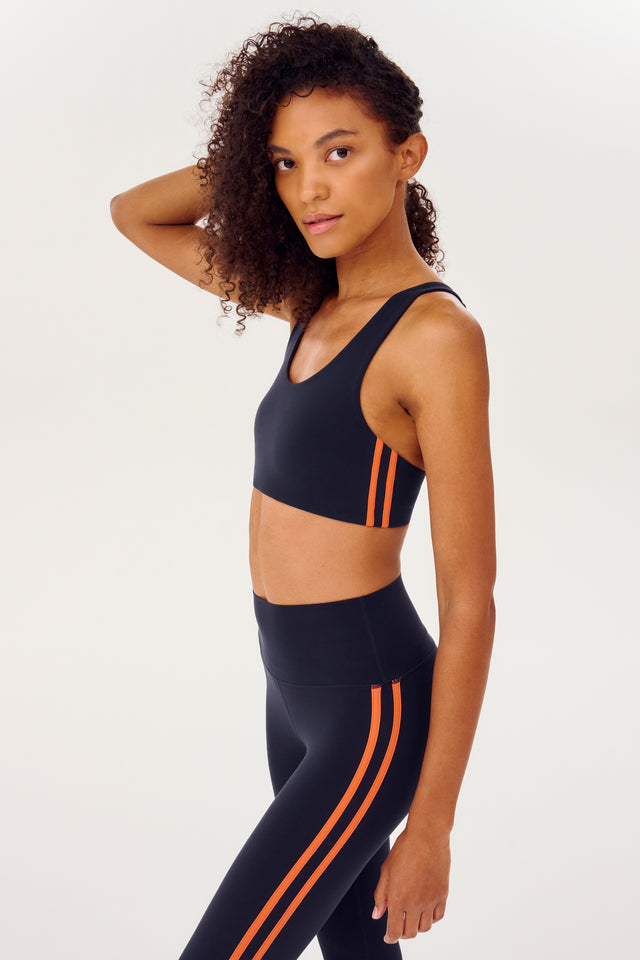 Side view of girl wearing dark blue sports bra with two thin red stripes down the side and dark blue leggings 