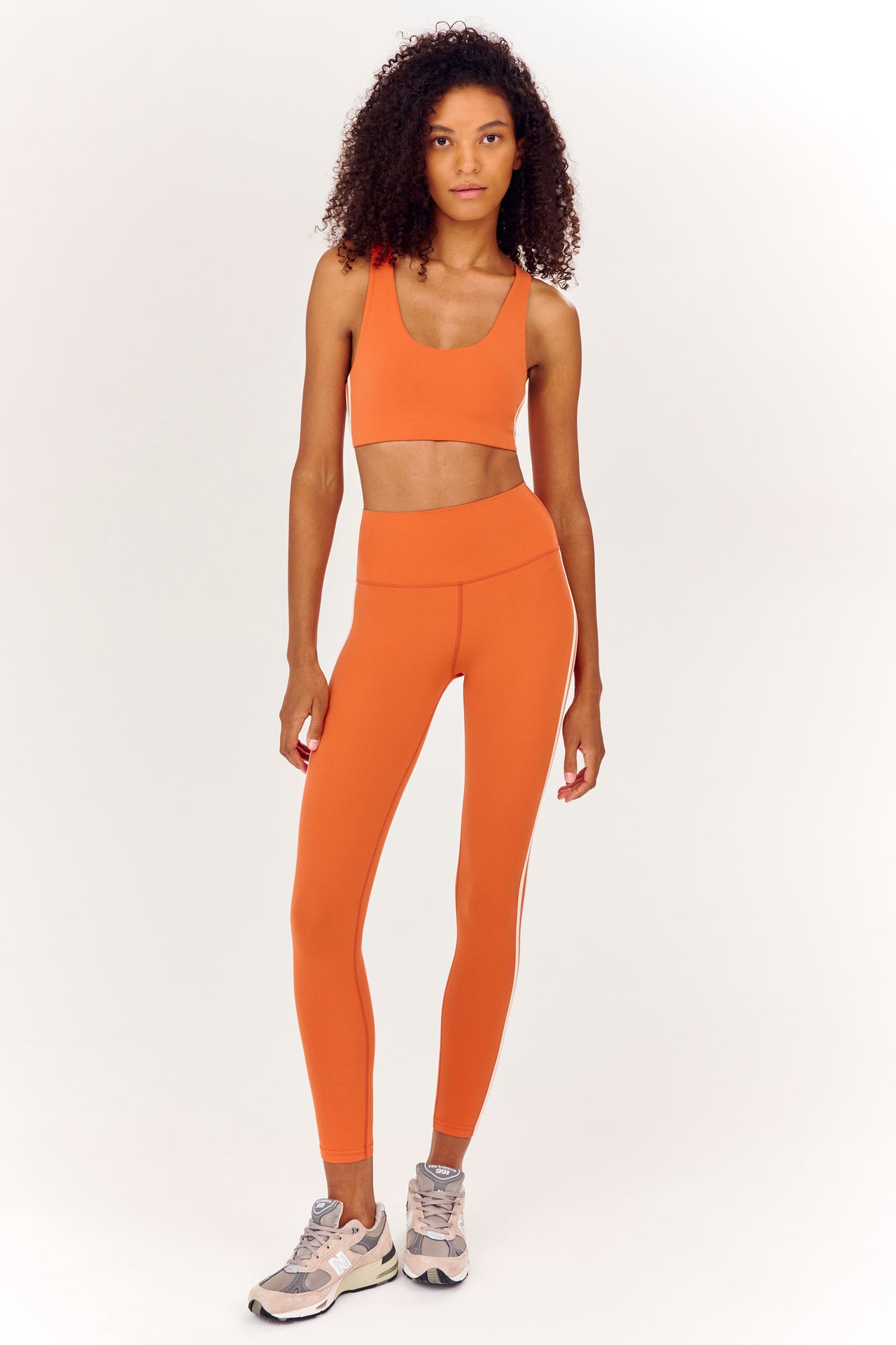 Full front view of girl wearing orange sports bra with two thin white stripes  down the side and orange leggings with white shoes