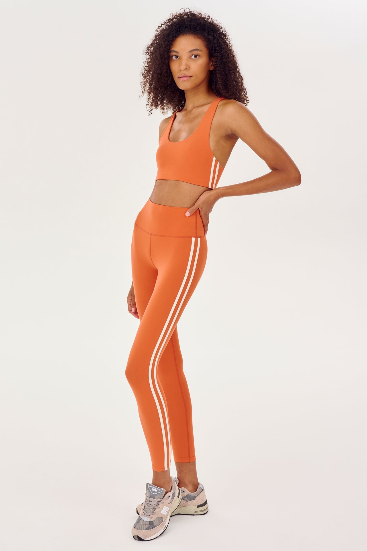 Full side view of girl wearing orange leggings with two thin white stripes down the side and a orange sports bra with white shoes