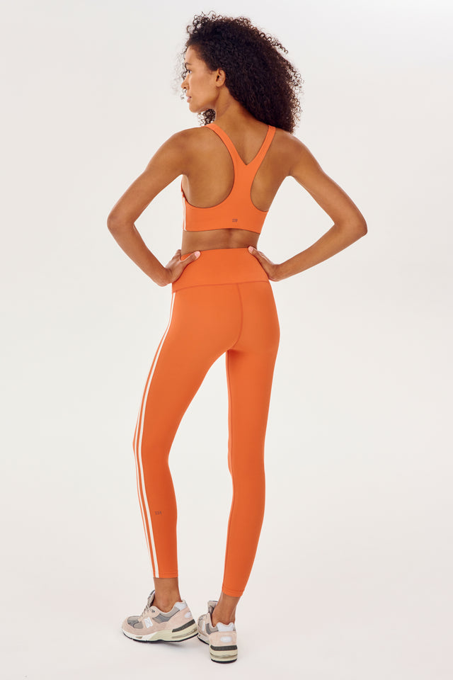 Full back view of girl wearing orange leggings with two thin white stripes down the side and a orange sports bra with white shoes