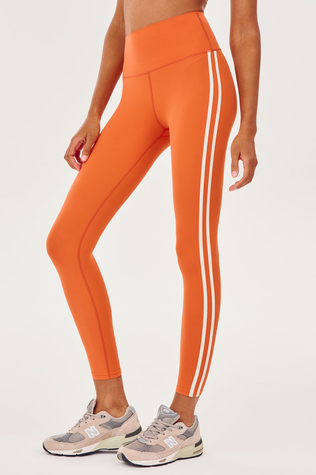 Side view of girl wearing orange leggings with two thin white stripes down the side with white shoes 