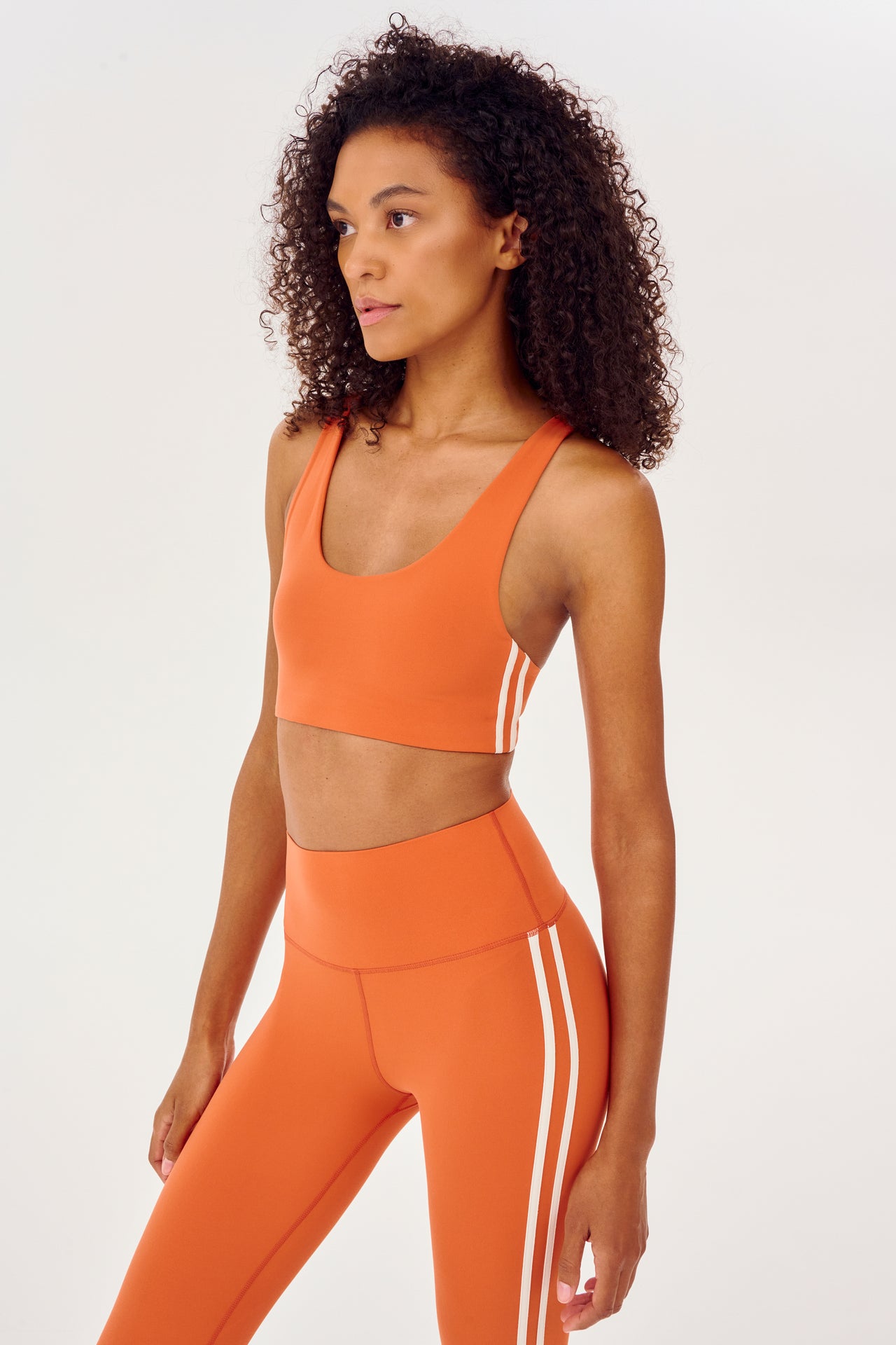 Side view of girl wearing orange sports bra with two thin white stripes  down the side and orange leggings 