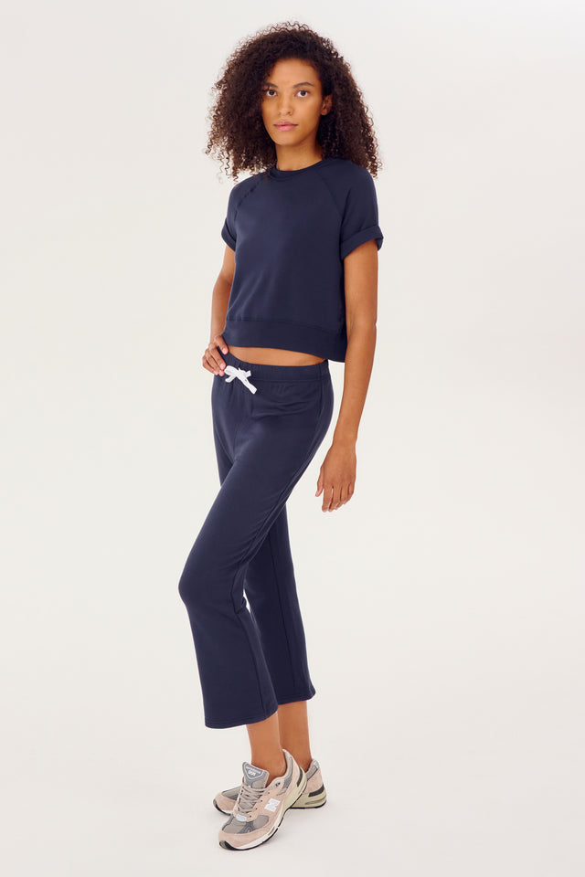 Full side view of girl wearing dark blue flared sweatpants that stop mid shin with a white string around waist with a cropped dark blue short sleeve shirt and multi colored shoes