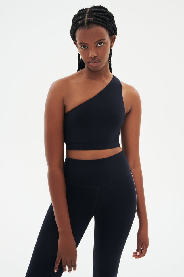 Front view of girl wearing a dark blue one shoulder bra and a black leggings with a white stripe down the side 