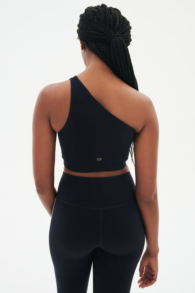 Back view of girl wearing a dark blue one shoulder bra and a black leggings with a white stripe down the side 