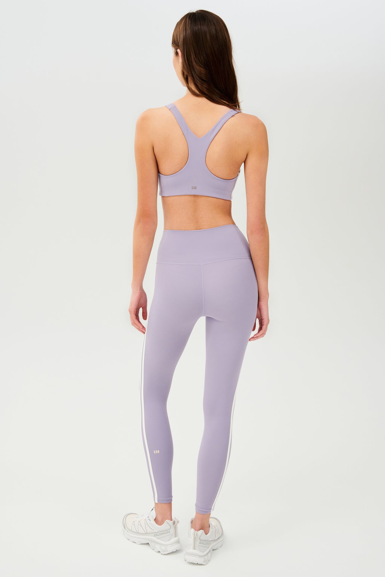 Full back view of girl wearing light purple leggings with two thin white stripes down the side and a light purple sports bra with white shoes