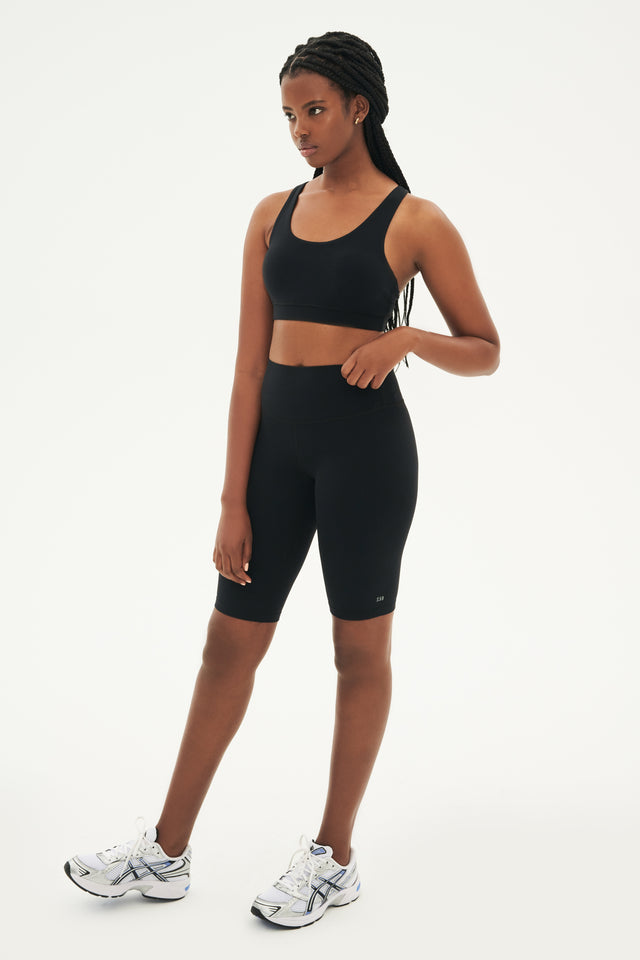 Full side view of girl wearing black sports bra and black high waisted bike shorts that stop right above the knee with white and grey shoes