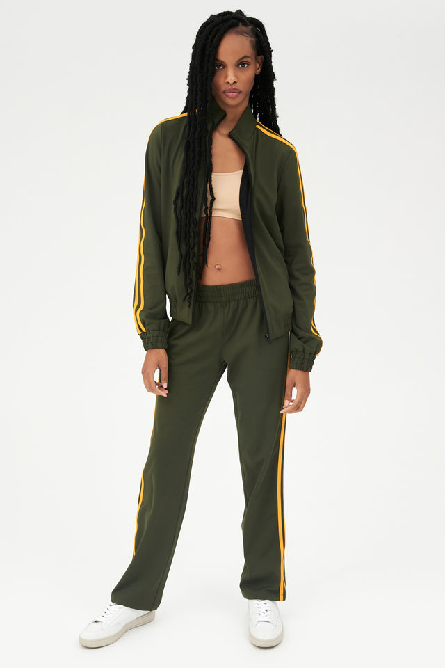Full front view of girl wearing dark green zip jacket that stops under chin with two yellow stripes down the side and a dark green sweatpants with white shoes