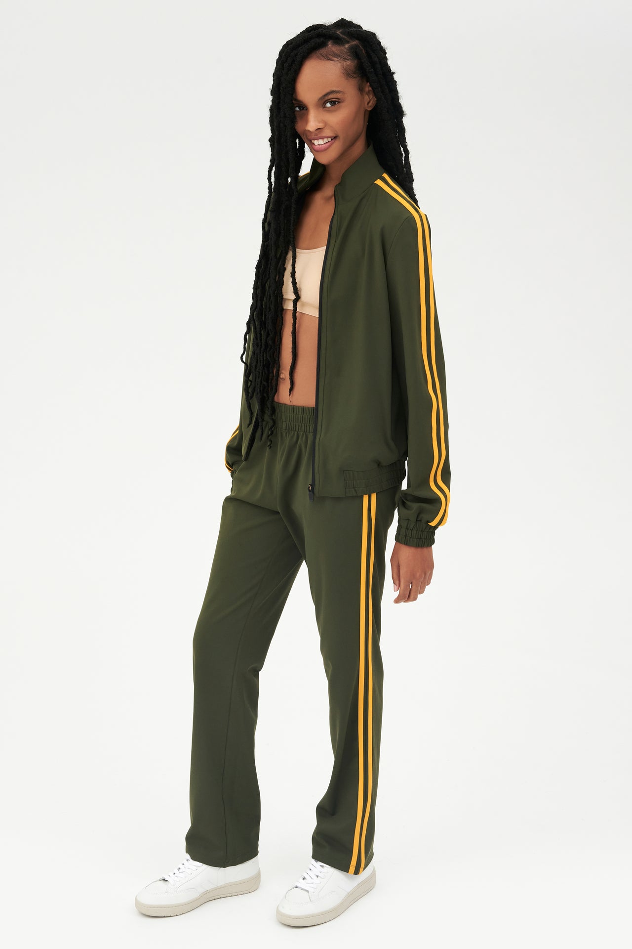 Full side view of girl wearing dark green zip jacket that stops under chin with two yellow stripes down the side and a dark green sweatpants with white shoes