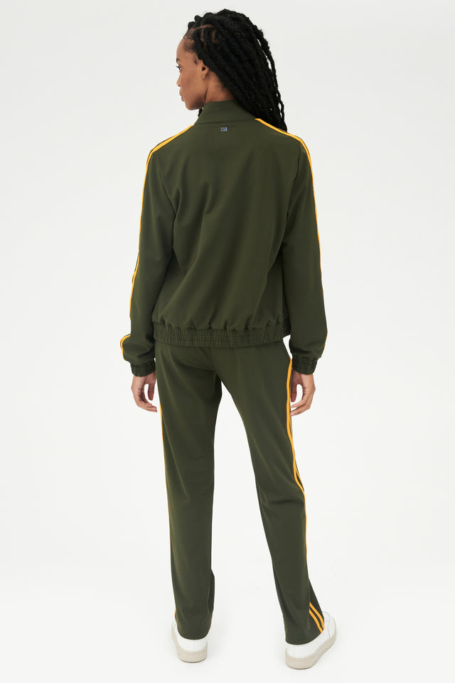 Full back view of girl wearing dark green zip jacket that stops under chin with two yellow stripes down the side and a dark green sweatpants with white shoes