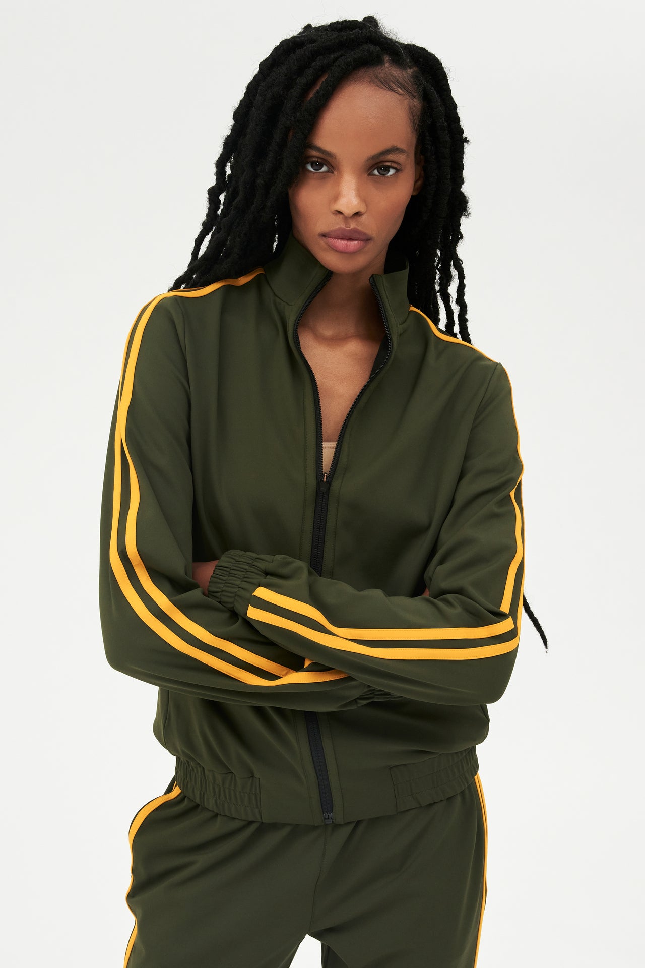 Front view of girl wearing dark green zip jacket that stops under chin with two yellow stripes down the side and a dark green sweatpants