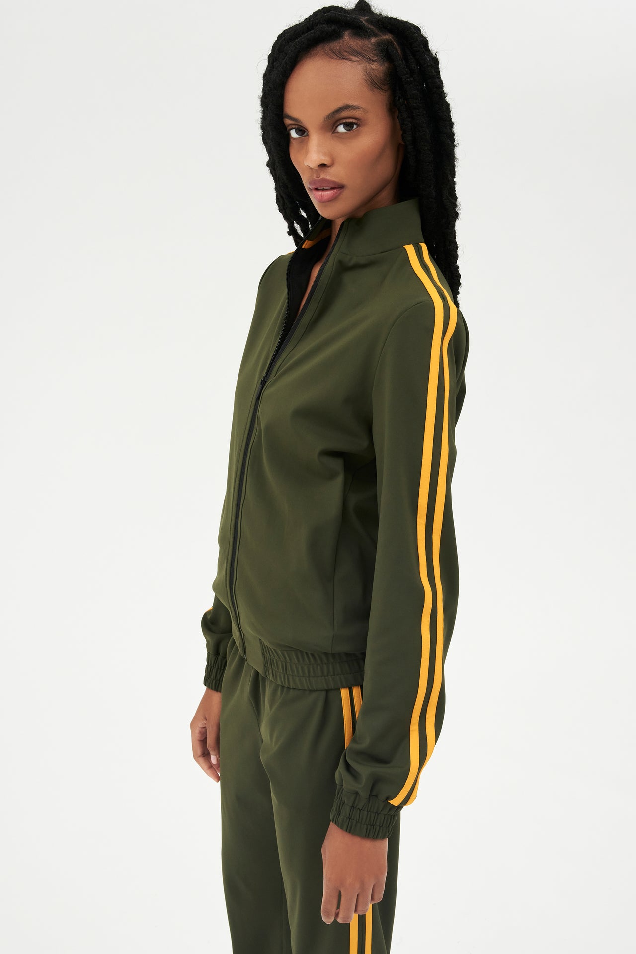 Side view of girl wearing dark green zip jacket that stops under chin with two yellow stripes down the side and a dark green sweatpants 