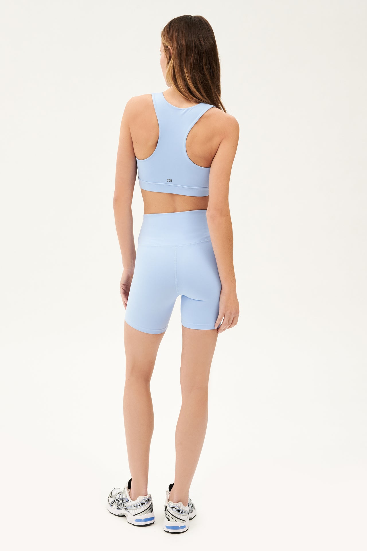 Full back view of woman with dark blonde straight hair wearing pale blue racer strap bra and pale blue high waist bike shorts paired with white shoes with black stripes