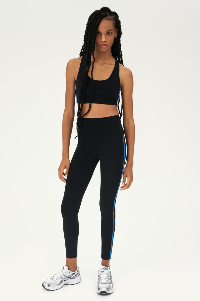 Full front view of girl wearing black sports bra with thin white and blue stripes down the side and black leggings with white shoes