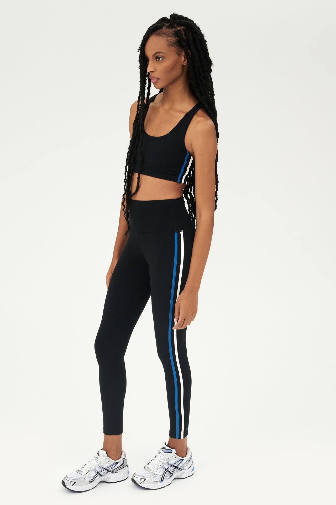 Full side view of girl wearing black sports bra with thin white and blue stripes down the side and black leggings with white shoes