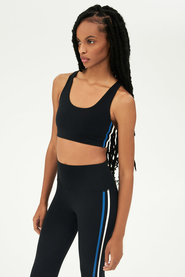 Side view of girl wearing black sports bra with thin white and blue stripes down the side and black leggings 