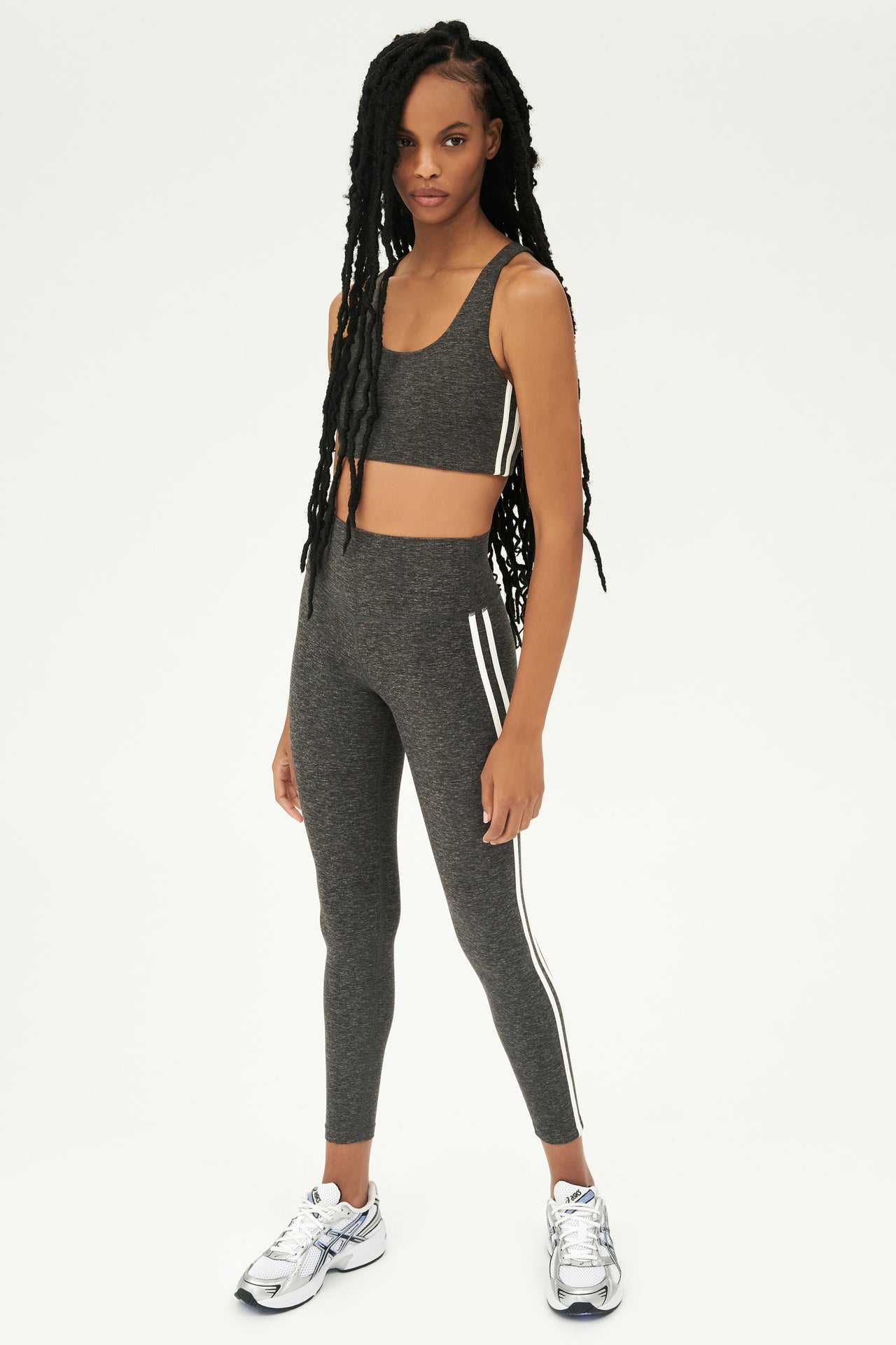 Full front view of girl wearing light dark grey sports bra with two thin white stripes down the side and light dark grey leggings with white shoes