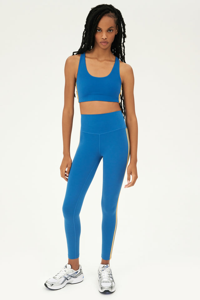 Full front view of girl wearing blue sports bra with two thin yellow stripes down the side and blue leggings with white shoes